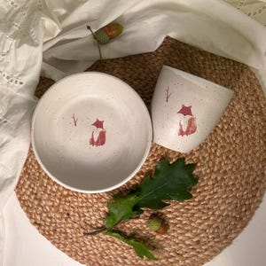 Könnte beinhalten: Eine gesprenkelte cremeweiße Keramikschale und ein passender Becher, beide mit einer kleinen handgemalten roten Fuchsdarstellung und einem dünnen roten Zweigmotiv. Das Set ist mit einem grünen Eichenblatt und zwei kleinen Eicheln arrangiert.