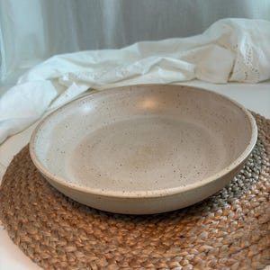 Könnte beinhalten: Eine flache, runde Keramikschale mit gesprenkelter beiger Oberfläche. Die Schale hat eine glatte, matte Textur mit kleinen dunkelbraunen Sprenkeln, die über das Innere und Äußere verteilt sind, sowie einen schlichten, runden Rand.