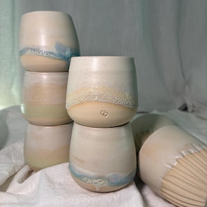 Könnte beinhalten: Sechs handgefertigte Keramikbecher. Die Becher sind cremefarben mit einer gesprenkelten Oberfläche. Einige haben eine blaue oder gelbe Glasur in Bodennähe. Ein Becher hat ein Herzsymbol. Die Becher stehen auf einem weißen Tuch.