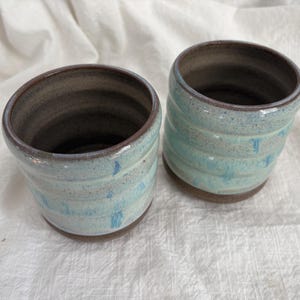 Könnte beinhalten: Zwei handgefertigte Keramikbecher mit hellblauer und brauner Glasur. Die Becher haben eine zylindrische Form mit horizontalen Rillen und einem dunkelbraunen Innenraum. Die Becher eignen sich für Kaffee, Tee oder andere Getränke.