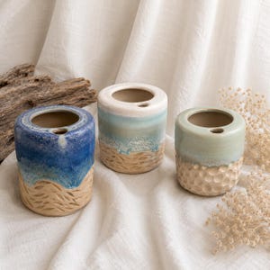Könnte beinhalten: Drei Keramik-Zahnbürstenhalter mit Küsten-Design. Jeder Halter ist zylindrisch mit strukturierter Basis und glatter, glasierter Oberseite. Die Farben umfassen Blau, Weiß und Beige, die ein Strandthema hervorrufen. Die Halter sind etwa 10 cm hoch.