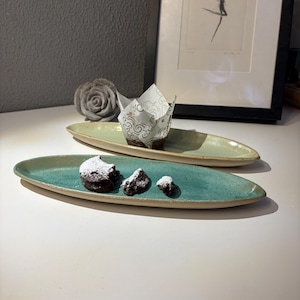 Könnte beinhalten: Zwei ovale Keramik-Servierplatten, eine türkisfarbene und eine hellgrüne, jeweils mit leicht erhöhtem Rand. Die türkisfarbene Platte enthält drei mit Puderzucker bestäubte Schokoladenstücke. Die grüne Platte enthält einen Muffin in einer dekorativen Papierform.