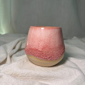 Könnte beinhalten: Ein Keramikbecher mit abgerundeter Form, der eine glatte, gesprenkelte rosa Glasur im oberen Bereich, ein strukturiertes, raues rosa Band in der Mitte und einen schlichten, matten beigen Boden aufweist, was einen deutlichen Kontrast erzeugt.