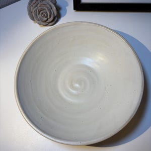 Könnte beinhalten: Eine handgefertigte, cremefarbene Keramikschale mit einem breiten, flachen Design. Die Schale hat eine leicht strukturierte Oberfläche und ein subtiles Spiralmuster in der Mitte. Ein kleines, graues, rosenförmiges Dekorationsobjekt befindet sich in der Nähe.