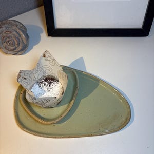 Könnte beinhalten: Ein hellgrünes Keramik-Serviertablett mit organischer Form. Ein kleineres, passendes Tablett enthält einen Schokoladenmuffin, der mit Puderzucker bestäubt ist. Der Muffin ist in gemustertes Papier gewickelt. Das Tablett ist ca. 25 cm lang.