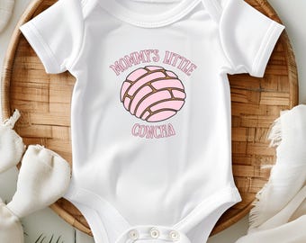 May include: White baby onesie with the text "Mommy's Little Concha" in pink lettering. The design features a pink concha bread illustration with brown lines. The onesie is short-sleeved and has snap closures.