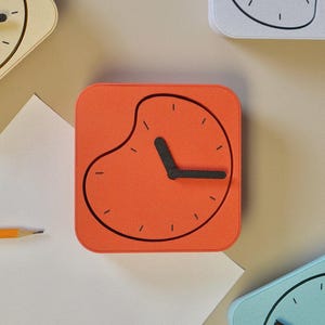 May include: Overhead view of several square clocks in different colours. The central clock is orange with a black clock face and hands. Other clocks are cream and blue. A pencil and paper are also in the image.