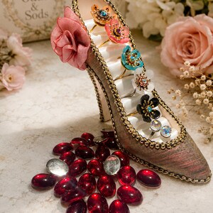 May include: A high-heeled shoe shaped ring holder with a fabric exterior, gold trim, and a pink fabric flower. The interior features white satin slots holding six floral and crystal rings. A collection of dark red oval glass cabochons lies below.