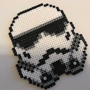 Puede incluir: Un diseño de píxeles en blanco y negro de un casco de Stormtrooper. El casco está hecho de pequeñas cuentas de plástico.
