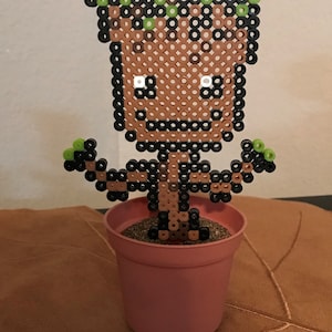 Peut inclure: Une figurine en pixel art marron et noir représentant un personnage de la série de films Les Gardiens de la Galaxie, Groot, dans un pot rose. La figurine a des feuilles vertes sur le dessus de la tête et se tient sur une surface brune.