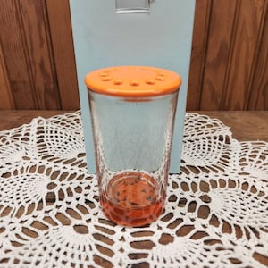 May include: A clear glass bottle with an orange lid and holes in the top. The bottle is sitting on a white crocheted doily.