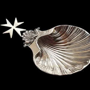 Peut inclure: Un plat en forme de coquillage argenté avec une étoile à huit branches décorative attachée. Le plat a une surface texturée et striée sur fond noir. L'étoile est argentée, brillante et réfléchissante.