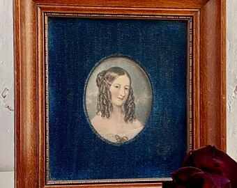 May include: A framed portrait of a woman with curly brown hair, wearing a white dress with a bow. The portrait is set against a blue velvet background and is framed in a dark wood frame.