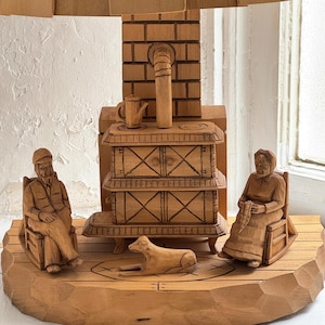 May include: A wooden folk art sculpture depicting a rustic scene. It features a stove, seated figures, and a dog. The figures are carved in detail, and the piece is made of natural wood.