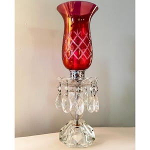 May include: A decorative candle holder with a red glass top featuring a diamond pattern. The holder has a silver base with crystal accents and hanging teardrop-shaped crystals. The base is made of clear, faceted glass.