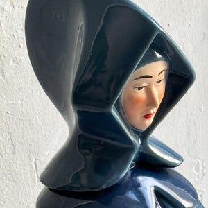 May include: A ceramic figurine of a woman wearing a blue hooded cloak. The woman's face is painted with delicate features and red lips.