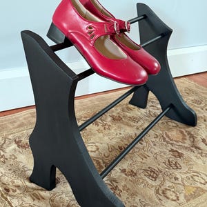 Peut inclure: Une paire de chaussures rouges de style Mary Jane avec une boucle et des d&eacute;tails d&eacute;coup&eacute;s, pos&eacute;e sur un pr&eacute;sentoir &agrave; chaussures noir. Le pr&eacute;sentoir a un design unique avec une base en forme de chaussure et des barres horizontales.
