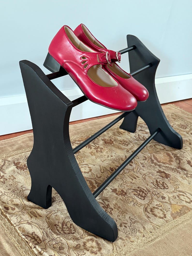 Peut inclure: Une paire de chaussures rouges de style Mary Jane avec une boucle et des d&eacute;tails d&eacute;coup&eacute;s, pos&eacute;e sur un pr&eacute;sentoir &agrave; chaussures noir. Le pr&eacute;sentoir a un design unique avec une base en forme de chaussure et des barres horizontales.