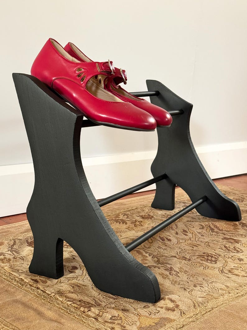 Peut inclure: Une paire de chaussures rouges de style Mary Jane repose sur un pr&eacute;sentoir &agrave; chaussures noir. Le pr&eacute;sentoir est en forme de talons hauts et poss&egrave;de une barre pour que les chaussures puissent reposer. Les chaussures ont des boucles et des d&eacute;coupes d&eacute;coratives.