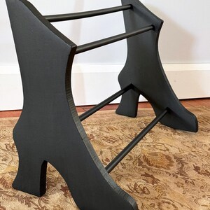 Peut inclure: Un porte-chaussures en bois noir avec un design de talon haut. Le support a deux supports verticaux en forme de talons hauts, reli&eacute;s par des barres horizontales. Le support est peint en noir et repose sur un tapis &agrave; motifs.