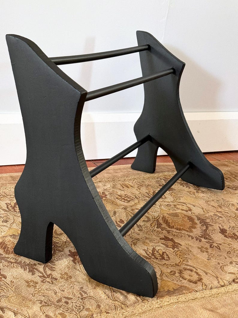 Peut inclure: Un porte-chaussures en bois noir avec un design de talon haut. Le support a deux supports verticaux en forme de talons hauts, reli&eacute;s par des barres horizontales. Le support est peint en noir et repose sur un tapis &agrave; motifs.