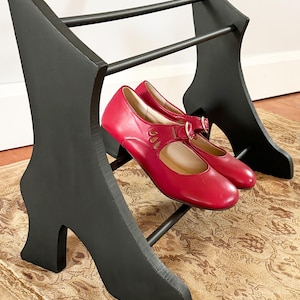 Peut inclure: Un pr&eacute;sentoir &agrave; chaussures noir en forme de talon haut, avec une paire de chaussures Mary Jane rouges. Le pr&eacute;sentoir est en bois et pr&eacute;sente un design unique. Les chaussures ont une boucle et des d&eacute;coupes d&eacute;coratives.