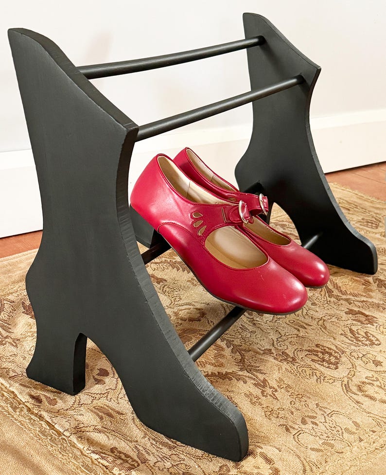 Peut inclure: Un pr&eacute;sentoir &agrave; chaussures noir en forme de talon haut, avec une paire de chaussures Mary Jane rouges. Le pr&eacute;sentoir est en bois et pr&eacute;sente un design unique. Les chaussures ont une boucle et des d&eacute;coupes d&eacute;coratives.