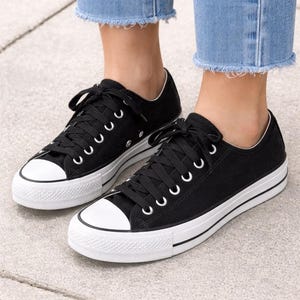 May include: Black canvas sneakers with white rubber soles and toe caps. The sneakers have black laces and white eyelets. The shoes are worn with blue jeans.