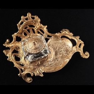 May include: A gold-toned metal inkwell stand featuring an ornate, swirling filigree frame with a clear, square glass ink container. The centre of the glass inkwell is topped with a circular, engraved metal lid that matches the base's finish.