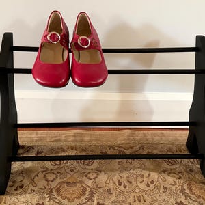 Peut inclure: Un pr&eacute;sentoir &agrave; chaussures en bois noir avec deux paires de chaussures rouges de style Mary Jane. Les chaussures ont une fermeture &agrave; boucle et sont expos&eacute;es sur un pr&eacute;sentoir &agrave; trois niveaux. Le pr&eacute;sentoir est sur un tapis &agrave; motifs.