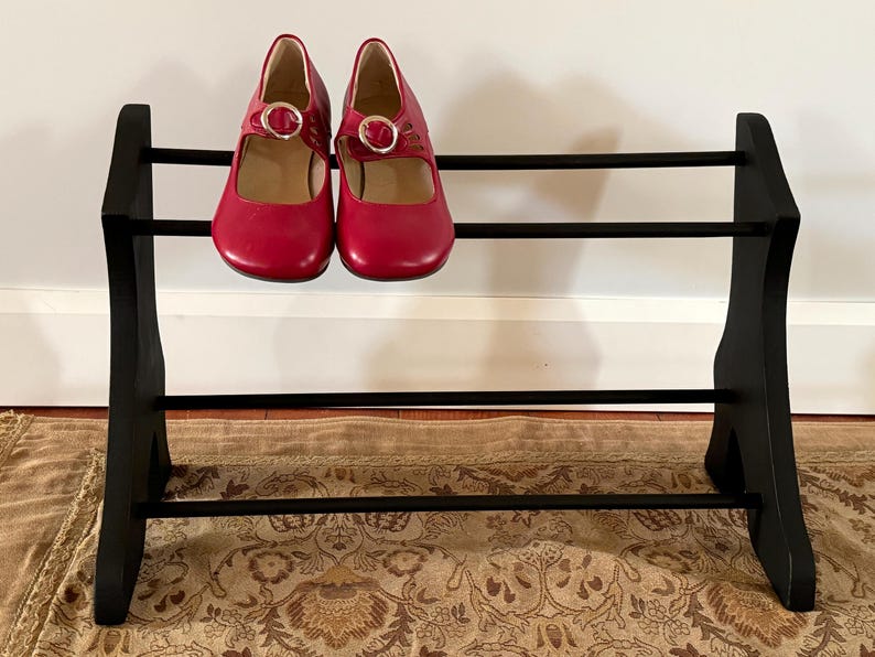 Peut inclure: Un pr&eacute;sentoir &agrave; chaussures en bois noir avec deux paires de chaussures rouges de style Mary Jane. Les chaussures ont une fermeture &agrave; boucle et sont expos&eacute;es sur un pr&eacute;sentoir &agrave; trois niveaux. Le pr&eacute;sentoir est sur un tapis &agrave; motifs.