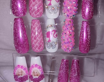 Könnte beinhalten: Set aus pinkfarbenen und weißen Press-on-Nägeln mit Glitzer, Strasssteinen und Barbie-Logos. Einige Nägel zeigen eine Barbie-Silhouette.