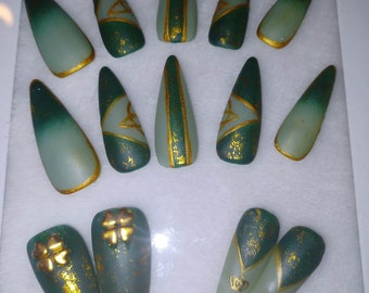 May include: A set of green and gold press-on nails. The nails feature various designs, including gold accents, Celtic knots, and shamrock patterns. The nails have a matte finish and are arranged in a white tray.
