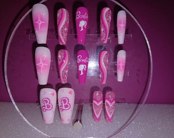 Peut inclure: Des ongles press-on roses et blancs sur le thème de Barbie. Certains ongles présentent un design ombré rose et blanc avec des paillettes, d'autres un design en spirale rose et blanc avec des paillettes, et certains un design en forme de cœur rose avec des paillettes. Le mot "Barbie" est écrit sur deux des ongles.