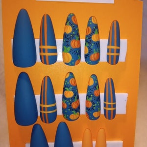 May include: A set of 12 blue and orange fake nails with pumpkin designs. The nails are arranged on a white foam board with the text "PUMPKIN PATCH" on a yellow background.