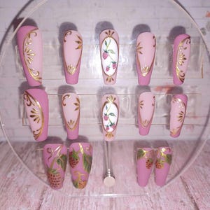 Puede incluir: Conjunto de uñas postizas rosas y doradas con un diseño floral. Las uñas presentan un óvalo blanco con un borde dorado y un diseño floral rosa con hojas verdes. Algunas uñas tienen un diseño de espiral dorado.