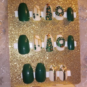 May include: Set of ten green and white Christmas-themed press-on nails. The nails feature a variety of designs, including Christmas trees, wreaths, and bows. The nails are displayed on a gold glitter background.
