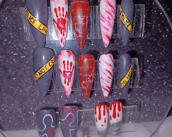 May include: Set of ten false nails with various Halloween-themed designs. The designs include a bloody handprint, a brick wall with blood splatters, a candy cane, a police line, and a ghost.