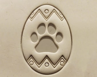 May include: A white clay egg with a paw print design. The egg is decorated with a simple geometric pattern.
