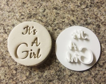 Puede incluir: Un cortador de galletas de plástico blanco con las palabras "It's A Girl" en una fuente cursiva. El cortador se muestra junto a una galleta que ha sido estampada con el cortador.