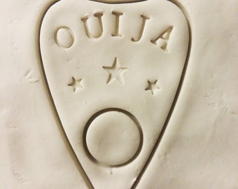May include: A white heart-shaped cutout with the word "OUIJA" in all caps and three stars above a circle.