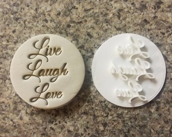 Puede incluir: Un cortador de galletas blanco con las palabras "Live Laugh Love" grabadas en él. El cortador se muestra con el lado grabado hacia arriba y el lado en blanco hacia abajo.