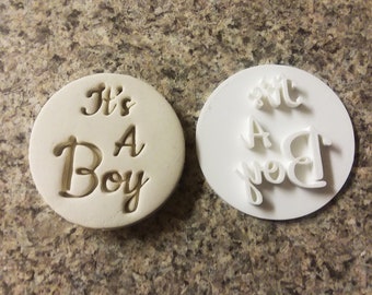 Puede incluir: Un cortador de galletas de plástico blanco con las palabras "It's A Boy" en una fuente cursiva. El cortador se muestra con las letras en relieve y las palabras están invertidas.
