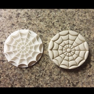 May include: Two white plastic cookie cutters shaped like spiderwebs. One cutter is flat and the other is raised.
