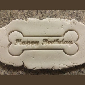 Puede incluir: Arcilla blanca con un cortador de galletas con forma de hueso presionado en &eacute;l. El cortador tiene las palabras "Happy Birthday" inscritas en &eacute;l.