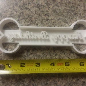 Puede incluir: Cortador de galletas de pl&aacute;stico blanco con forma de hueso con las palabras "Happy Birthday" en cursiva. El cortador mide 7,6 cm de largo.
