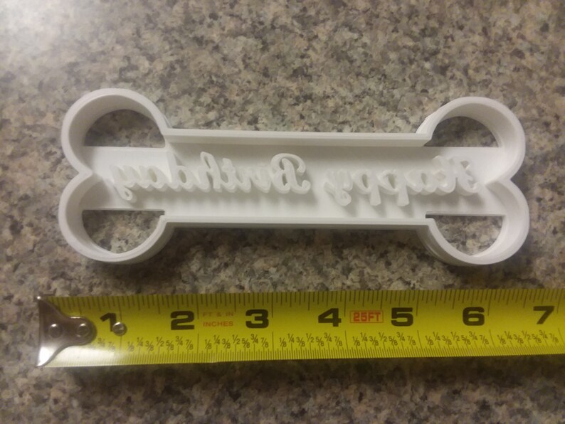 Puede incluir: Cortador de galletas de pl&aacute;stico blanco con forma de hueso con las palabras "Happy Birthday" en cursiva. El cortador mide 7,6 cm de largo.