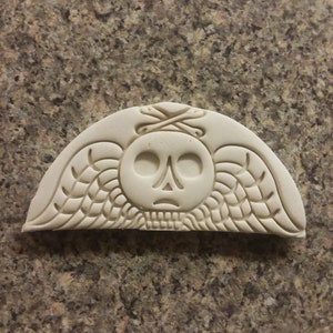 May include: White ceramic mold with a skull design and wings. The skull has crossed bones above it.