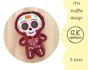 Puede incluir: Juguete de peluche bordado en forma de esqueleto con una cara de calavera de azúcar. El juguete es burdeos con detalles blancos y negros. El juguete está sobre una superficie de madera. El texto "ITH stuffie design" y "GK applique" son visibles en la imagen.