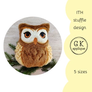May include: A brown and beige plush owl with orange eyes. The owl is sitting on a wooden surface with green pine branches.